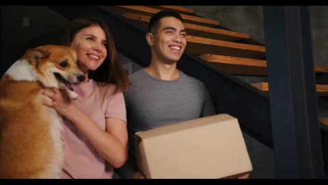 Smiling young man with box and woman with fluffy corgi dog in arms come into new spacious house