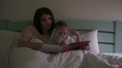 A mother trying to put her little girl to sleep by reading a storybook..