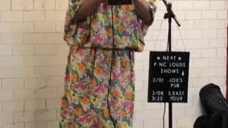 Man dressed up in makeup, wig, and large floral dress performs and sings in a subway station