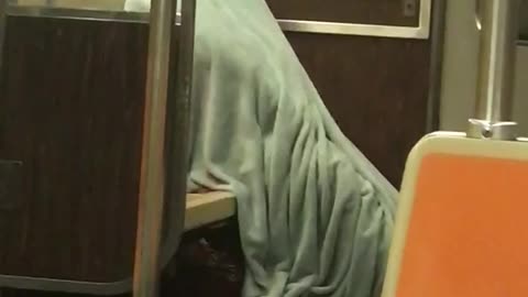 Person entirely covered from head to toe in gray blanket on subway train