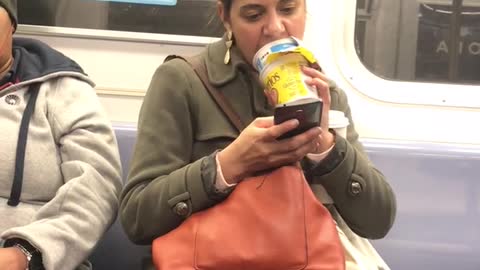 Woman uses her tongue to scoop cereal out of packaged cereal bowl on subway train