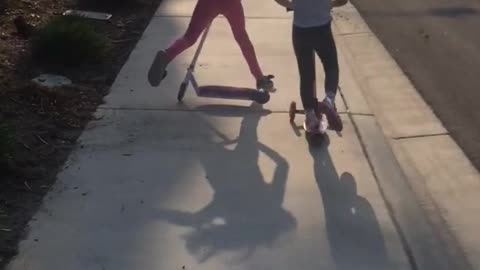 Two sisters racing on scooters, sister in front falls and trips other sister