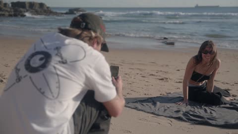 A man captures a smiling woman during a casual beach photo shoot against the ocean backdrop.