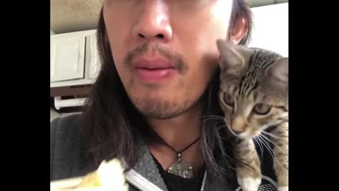 Cat sits on his owner's shoulder while he eats food