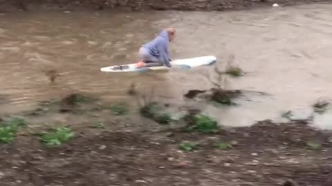 Girl rides surfboard down rain filled drainage ditch