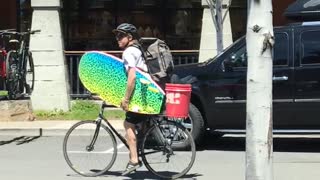 Guy riding bike holding colorful blue yellow surfboard