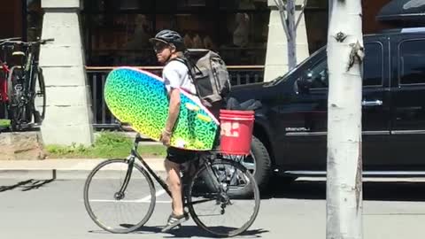 Guy riding bike holding colorful blue yellow surfboard