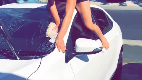 Girl washing car while sitting on top of windshield and falls