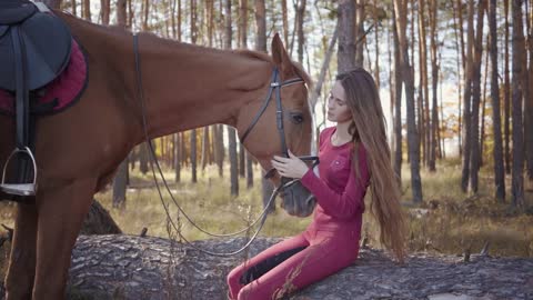 Portrait of young Caucasian woman sitting in the autumn forest and caressing horse's face