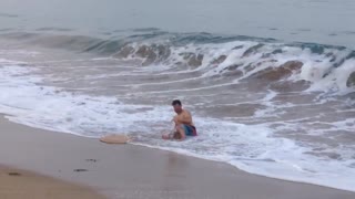 Guy runs to water on beach but trips over his board before he can step on it