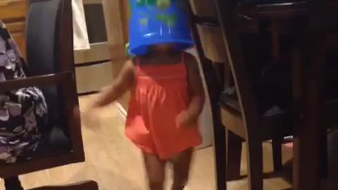 Little girl in dress with blue bucket on head walking around house runs into table